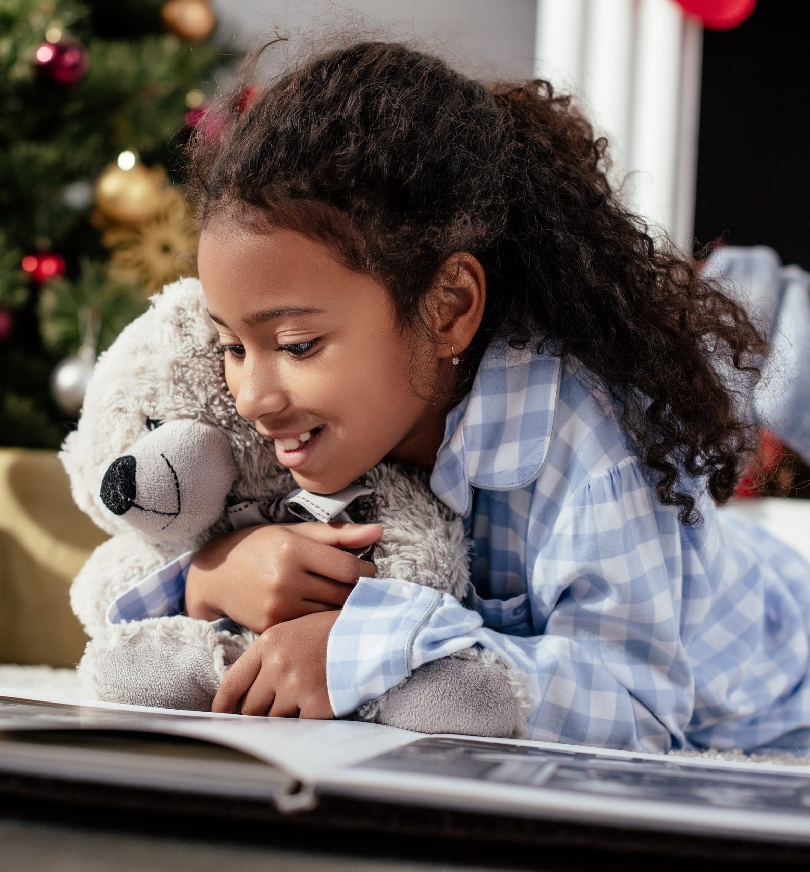 surprised and happy girl reading a book holding a teddy bear in front of christmas tree