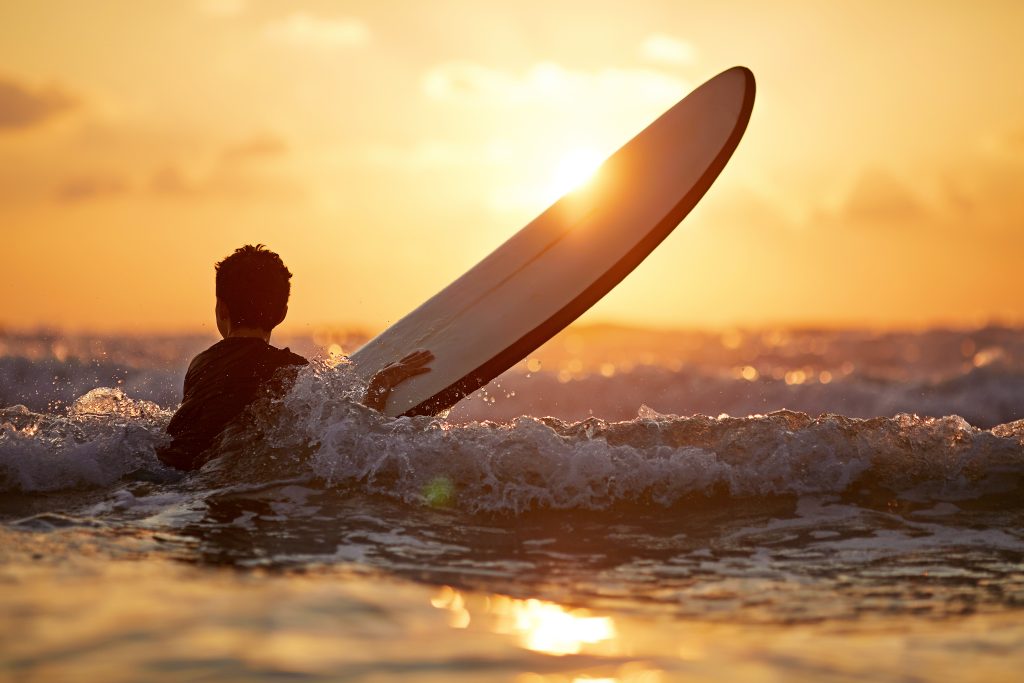 Back view of fearless kid floating surfboard at ocean with waves on sunny evening