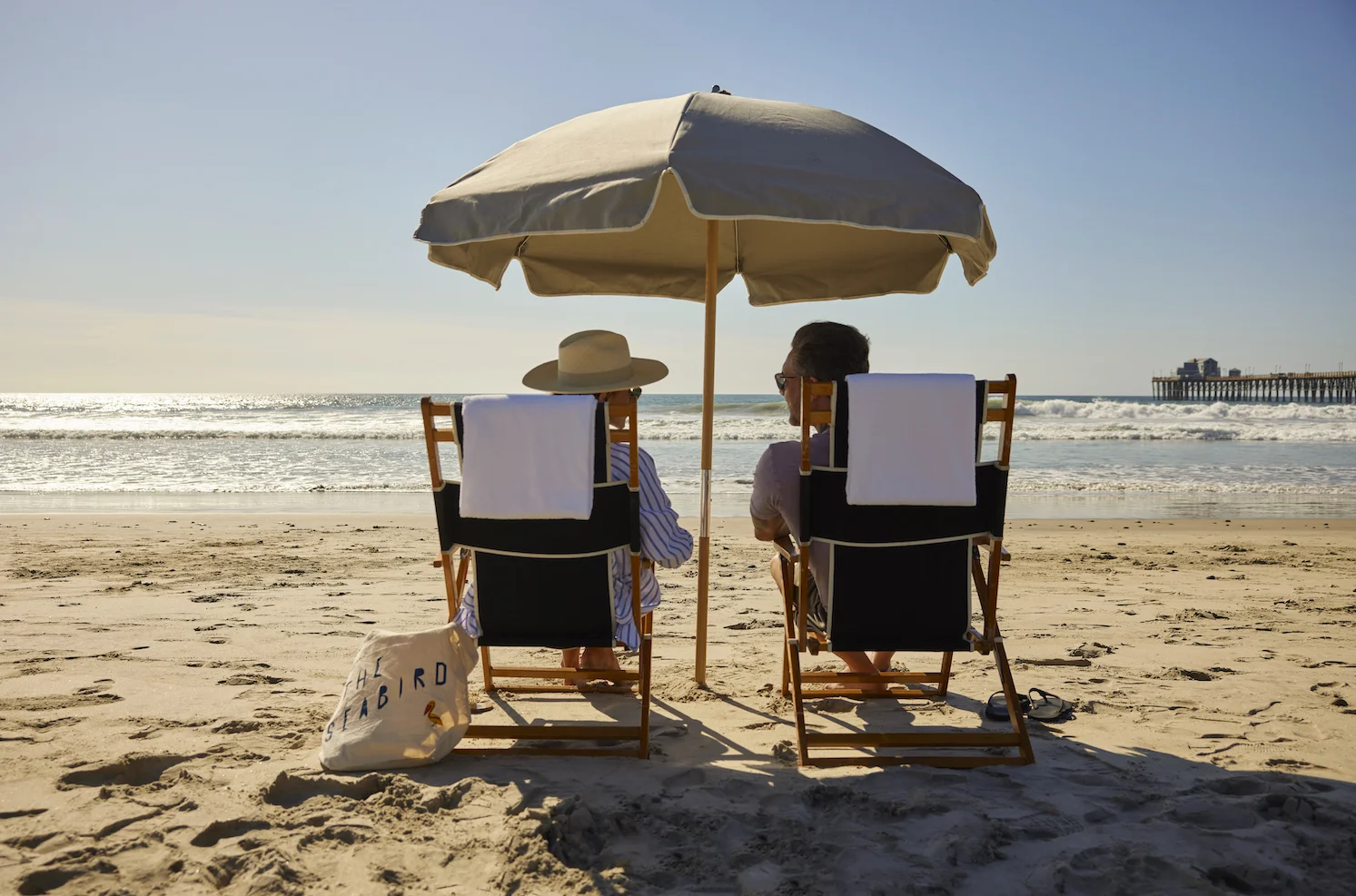 Two people relaxing in beach chairs under umbrella facing ocean with pier in distance