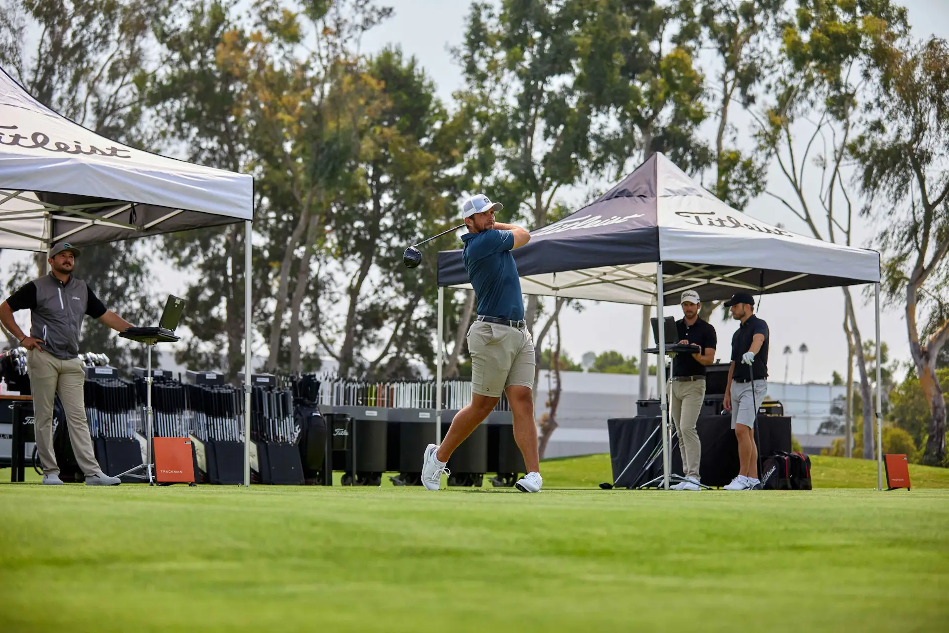 Golfer follows through after swing at outdoor equipment event, Titleist tents, staff, and trees in background.