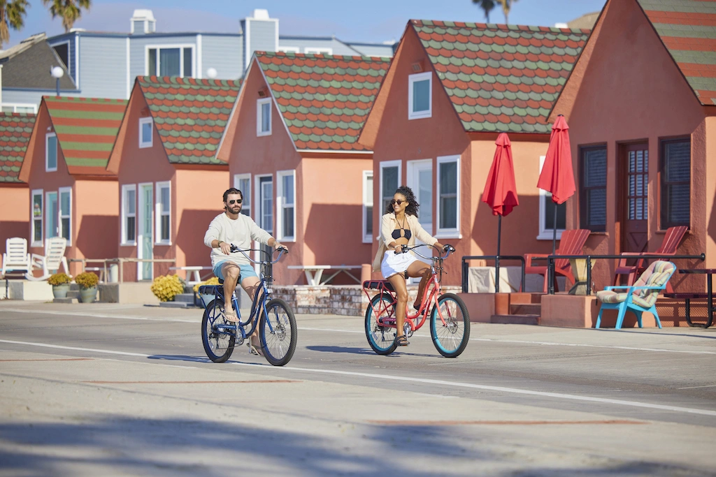 Couple riding bicycles along a sunny beachfront neighborhood with colorful cottages.
