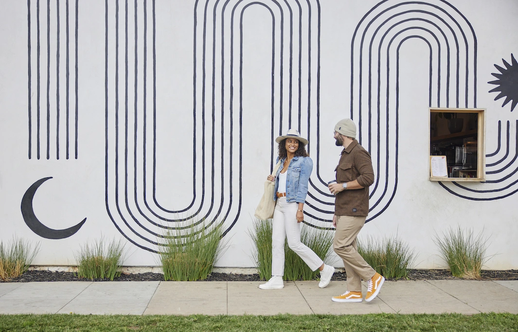 Two people walking past a modern mural wall, chatting and holding coffee.