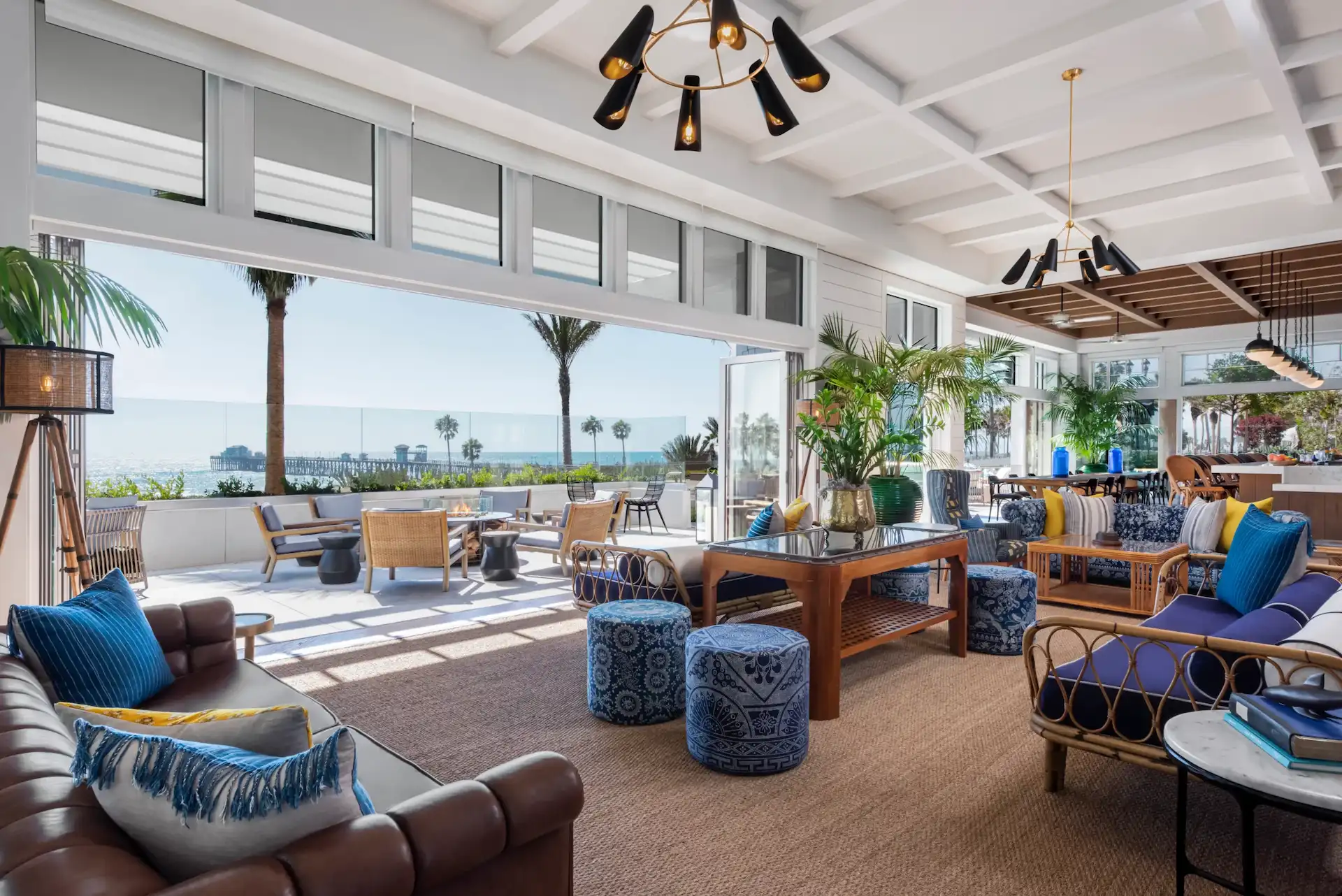 Indoor-outdoor lounge with tufted leather seating, woven textures, and ocean views framed by palm trees and breezy patio doors.