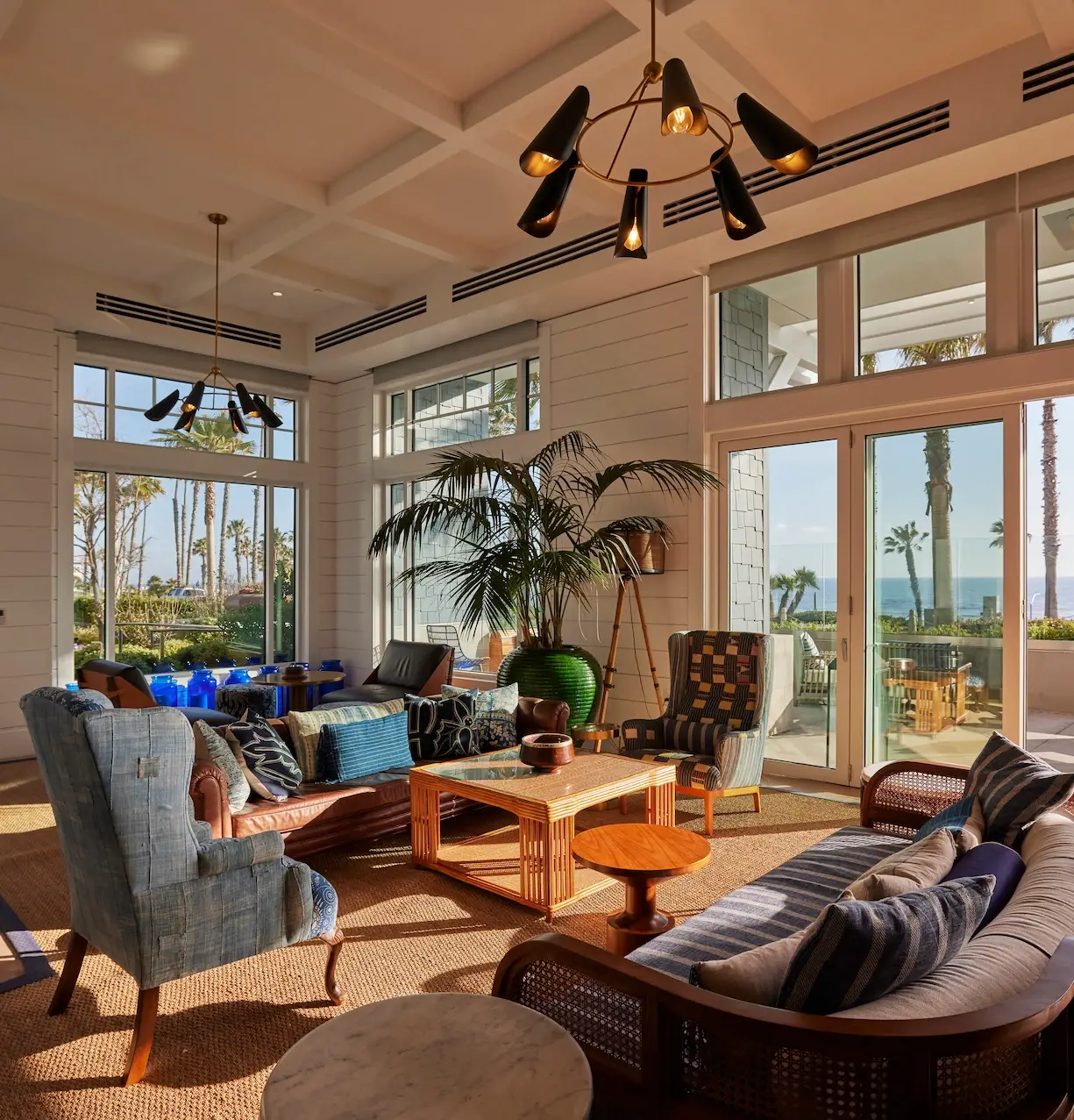 Coastal-style living room with striped sofa, armchairs, chandeliers, and large windows overlooking palm trees and ocean.