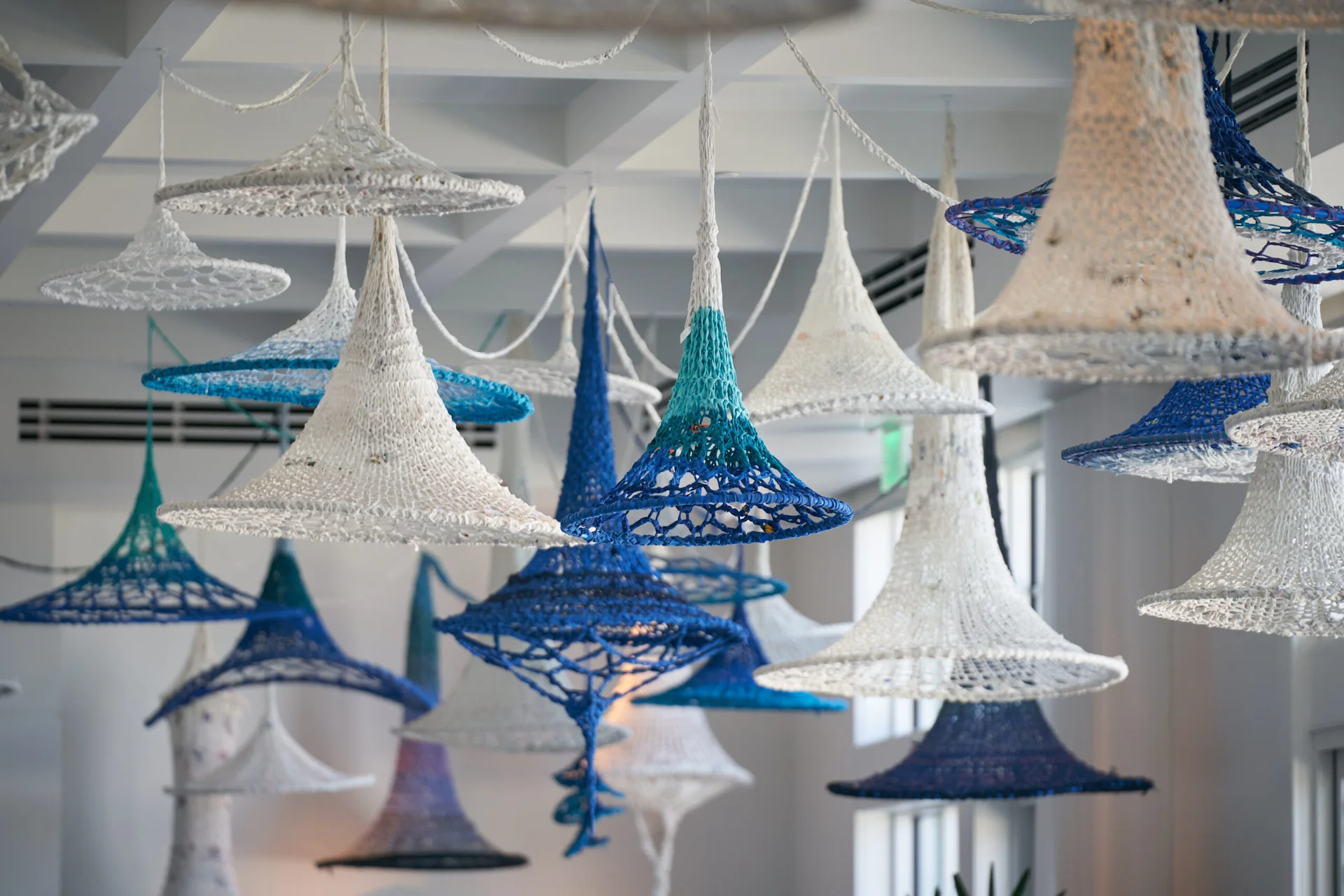 Hanging woven sculptures in white, blue, and turquoise yarn, suspended at varying heights in bright modern gallery space.