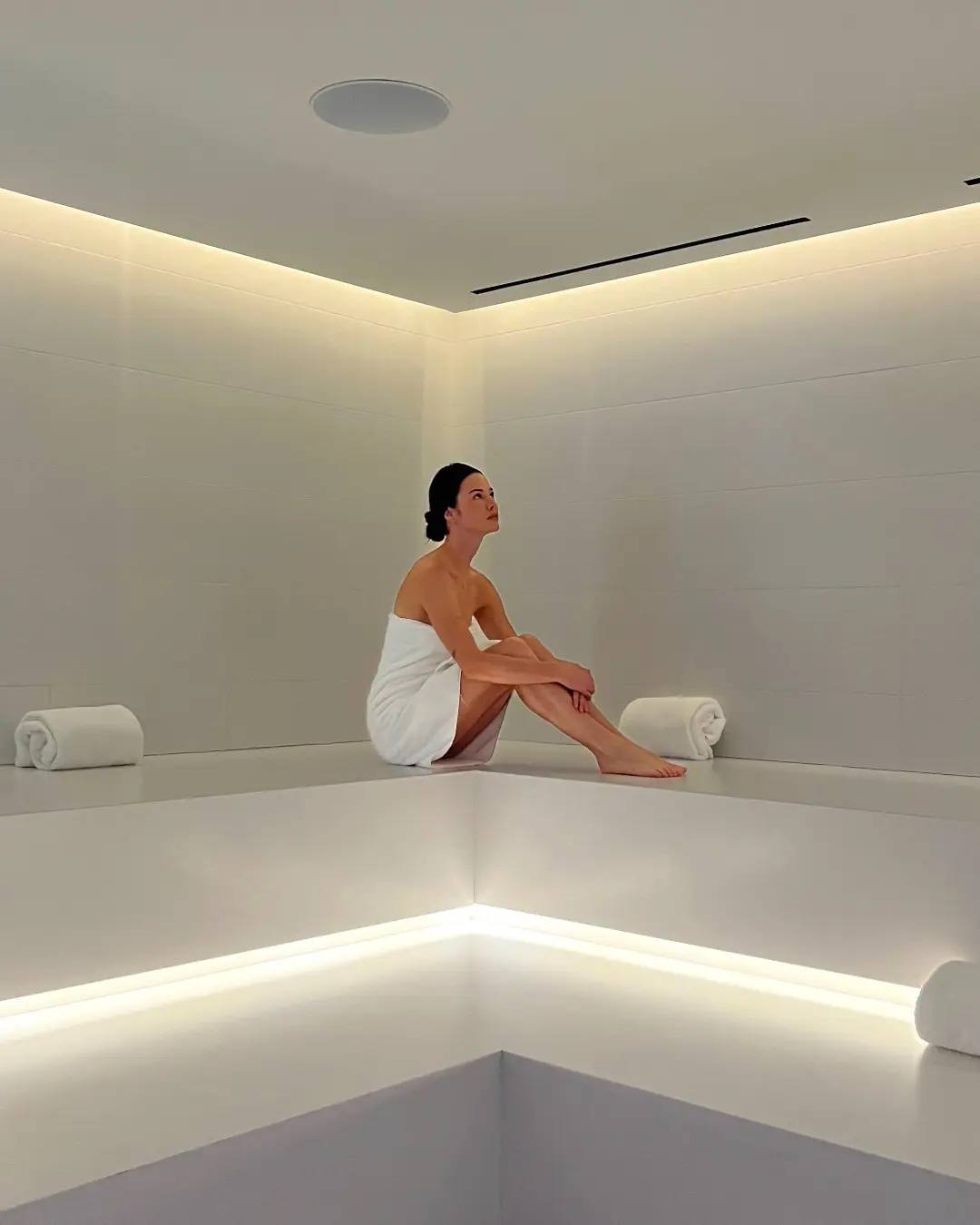 Woman in white towel sits in minimalist, modern spa room with soft lighting and rolled towels.