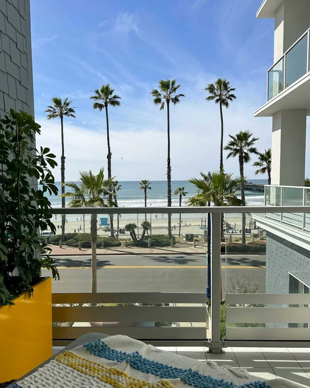Balcony view of sunny beach, palm trees, ocean waves, blue sky, and nearby modern white buildings.