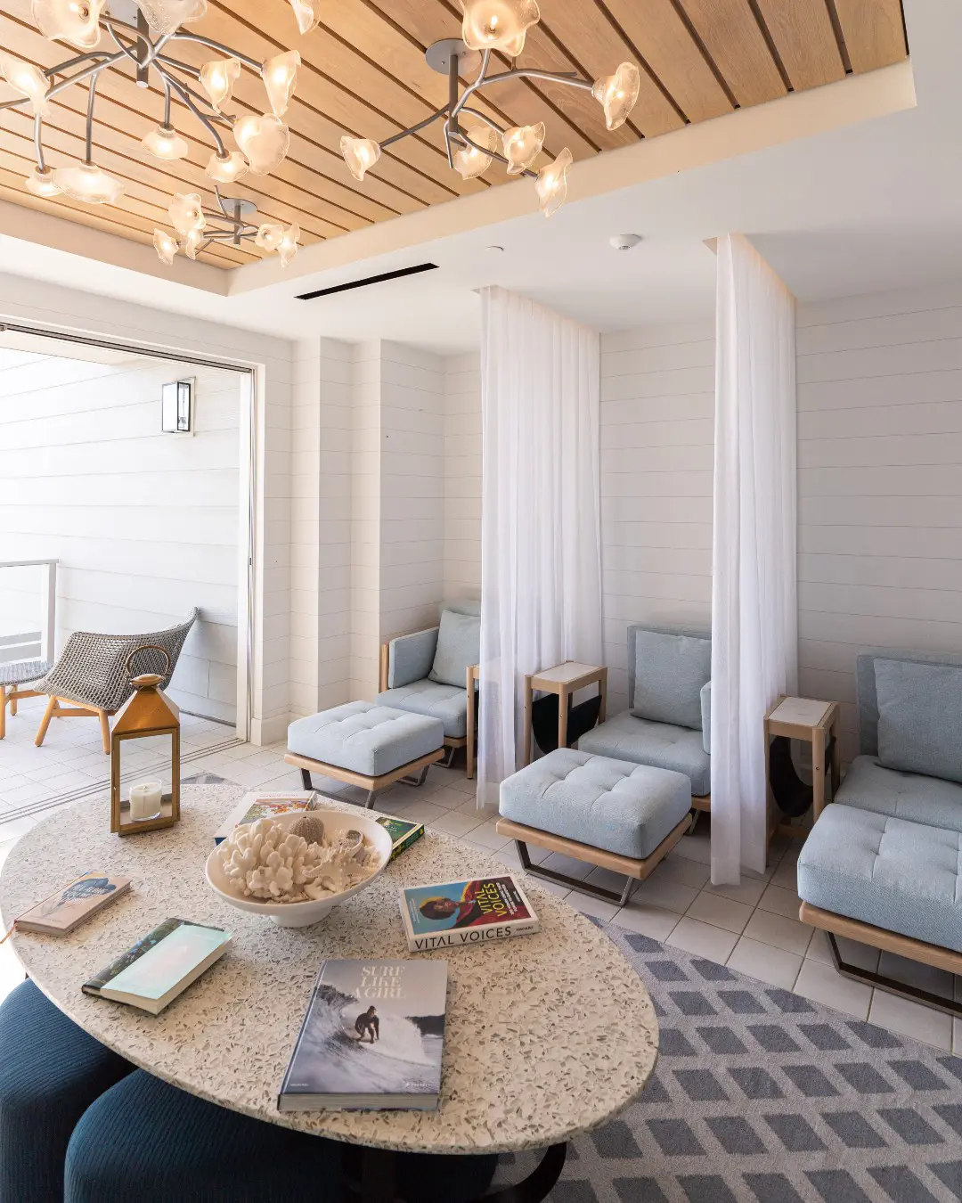 Bright spa lounge with soft seating, decorative lighting, books, coral centerpiece, and sheer white curtains accents.