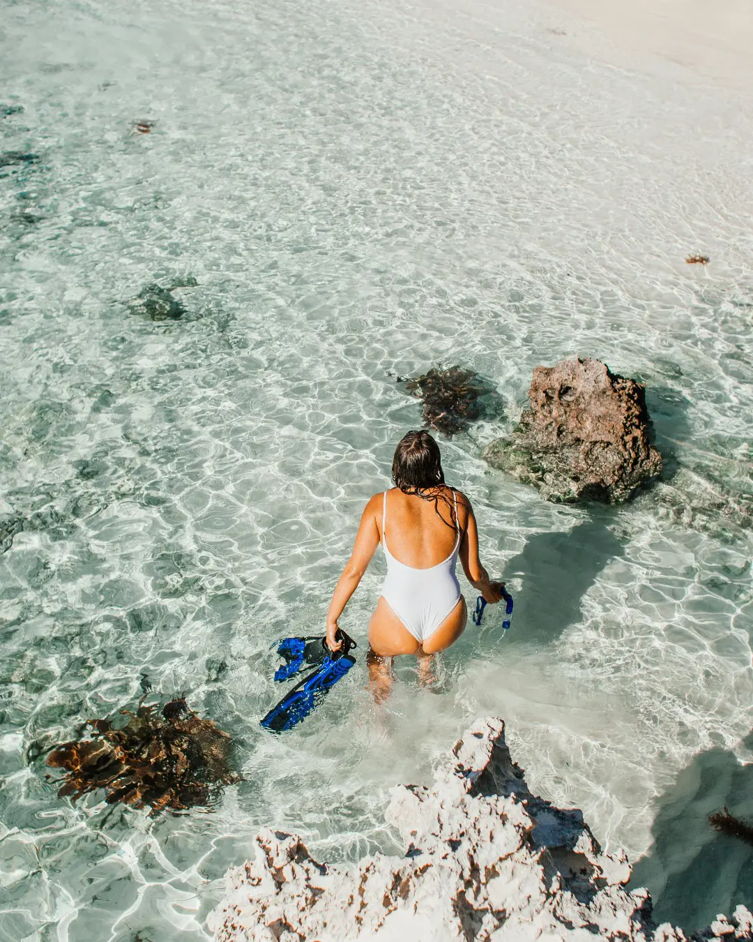 Woman in white swimsuit steps into clear shallow water, holding blue snorkel gear by rocky shore.