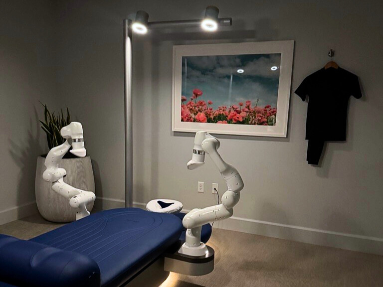 Robotic massage arms positioned over blue massage table in modern, softly lit therapy room with flower artwork.