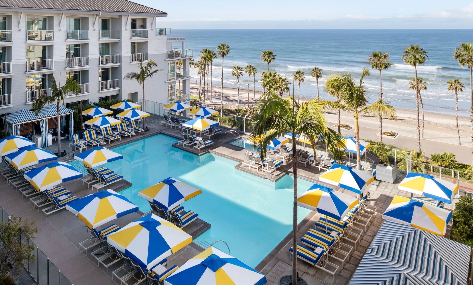 Beachfront resort with striped umbrellas, ocean-facing balconies, and a central pool framed by palm trees under clear blue skies.