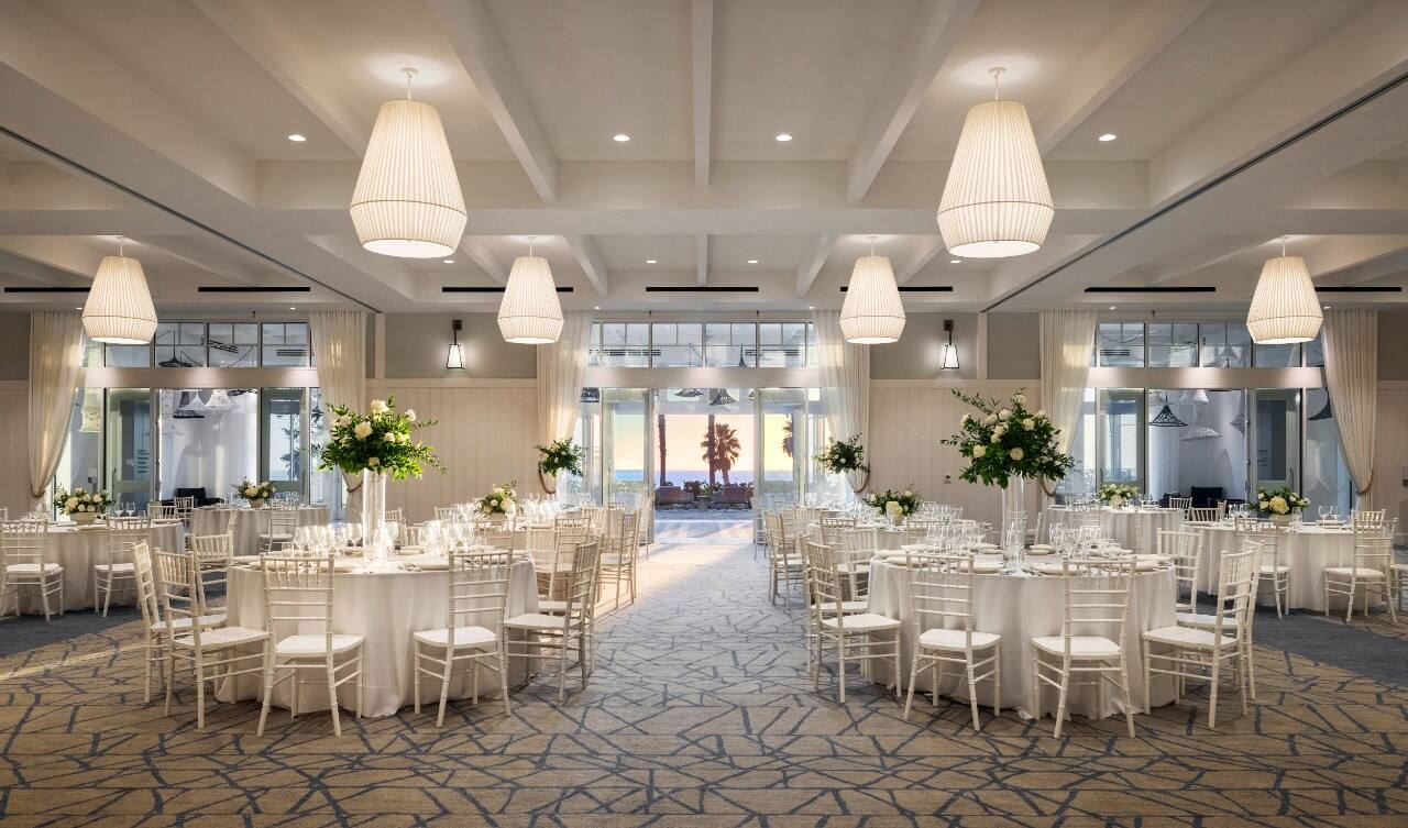 Bright wedding reception hall with round tables, white linens, tall floral centerpieces, and ocean view at sunset