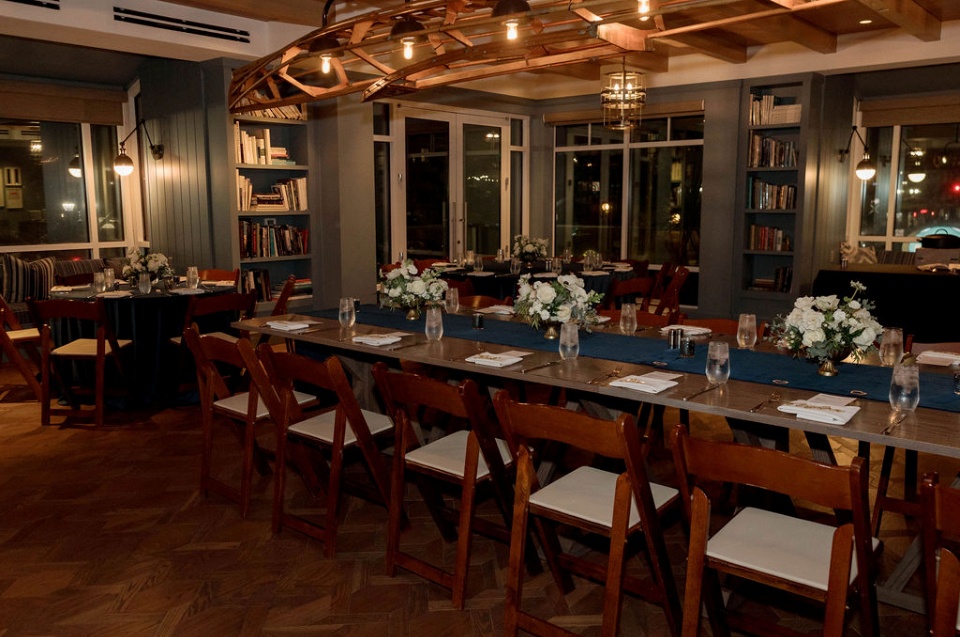 Warmly lit dining room with long wooden table, white floral centerpieces, bookshelves, and large windows at night