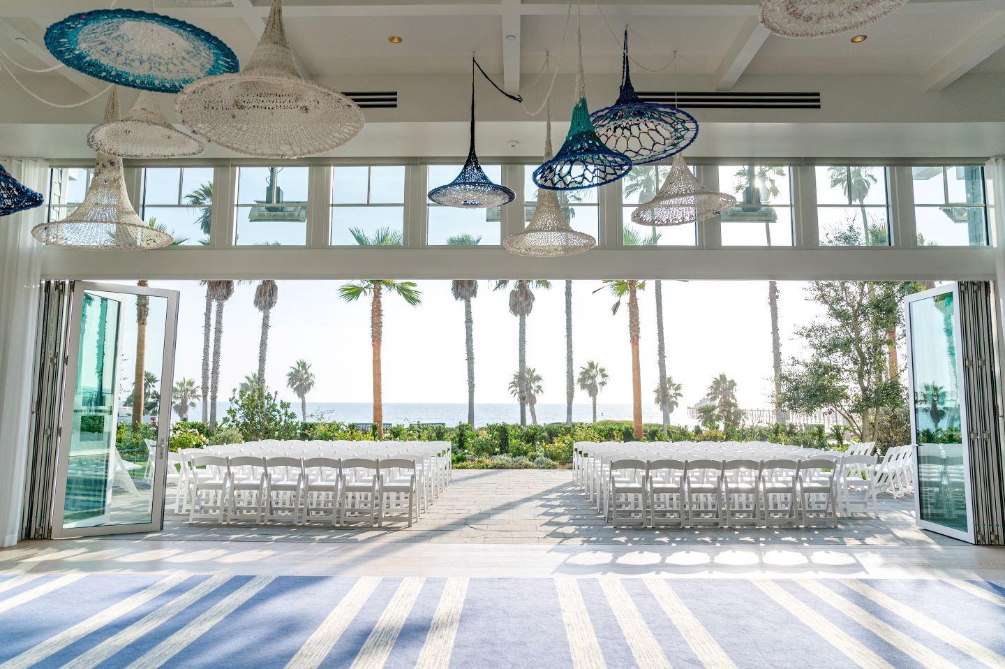 Indoor-outdoor wedding venue with ocean view, palm trees, white chairs, and woven hanging light fixtures