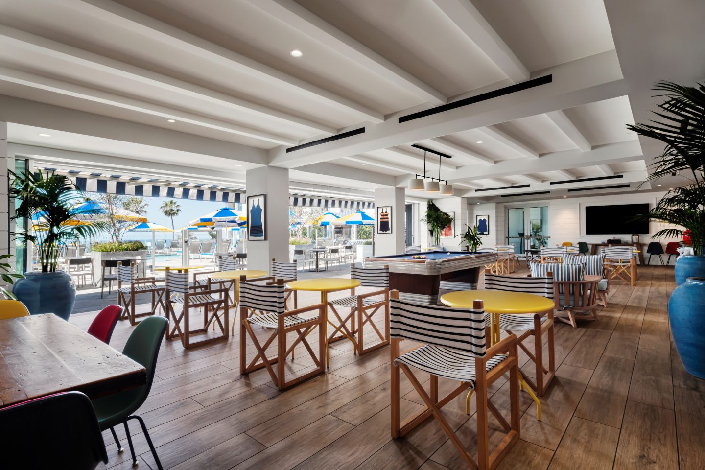 Bright poolside lounge with striped chairs, yellow tables, pool table, and blue umbrellas overlooking palm trees and ocean.