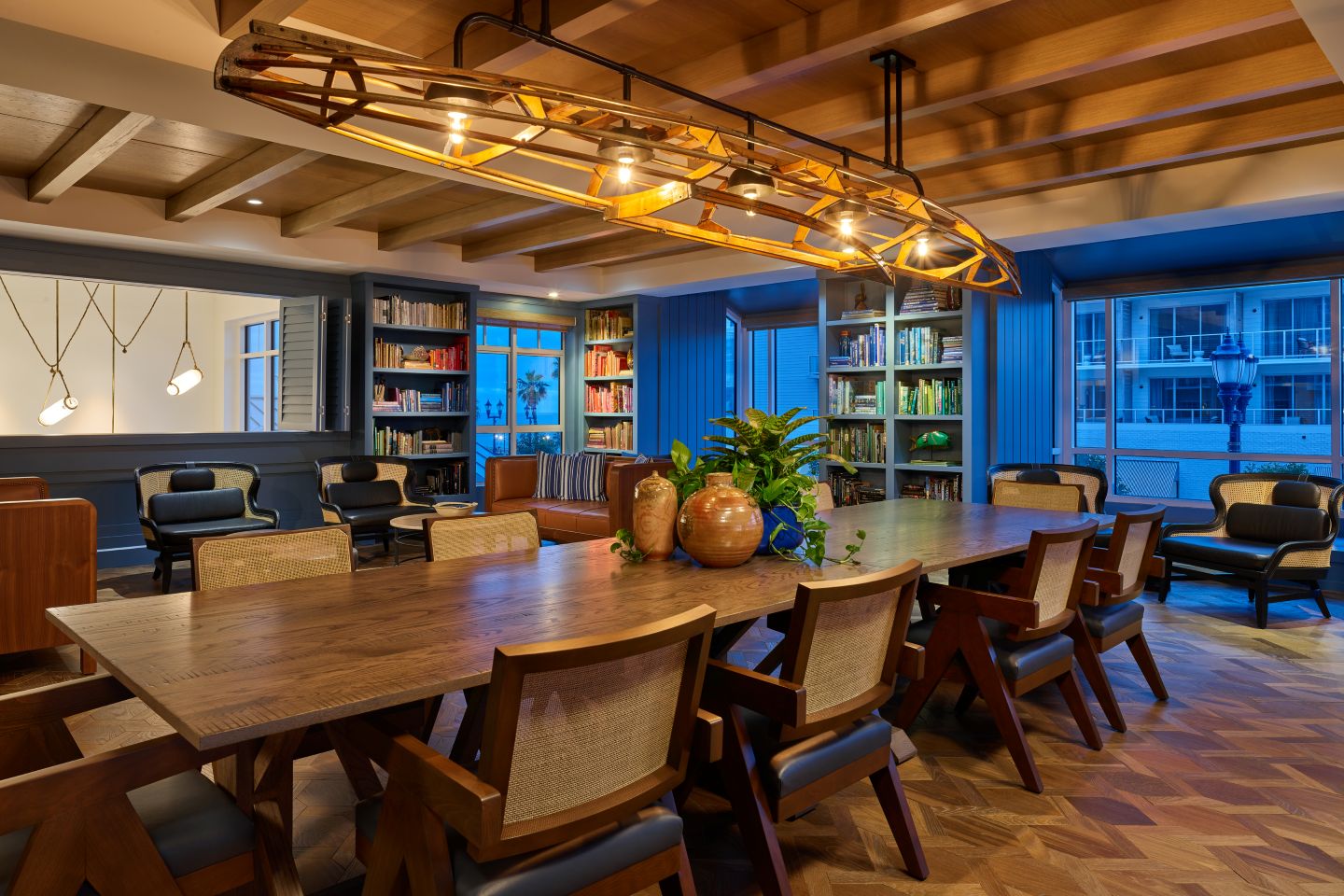 Warm, modern library-style dining room with long wooden table, woven chairs, bookshelves, and canoe light fixture