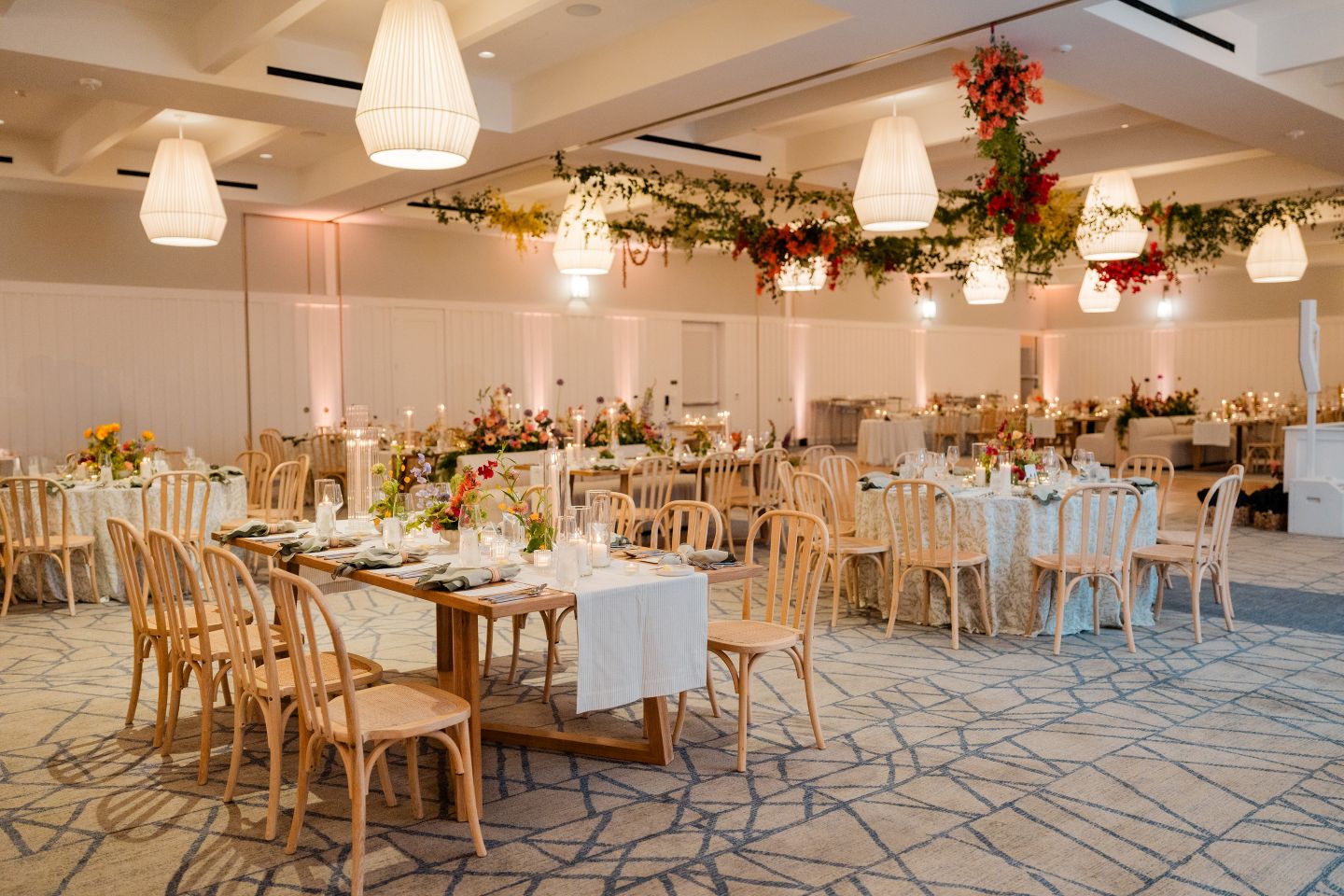Elegant wedding reception hall with round tables, floral centerpieces, hanging greenery, and warm ambient lighting