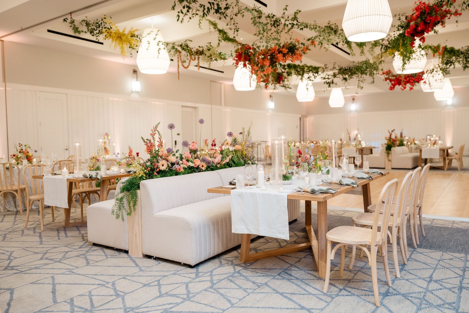 Elegant event space with wooden tables, floral centerpieces, cream seating, and hanging greenery with pendant lights