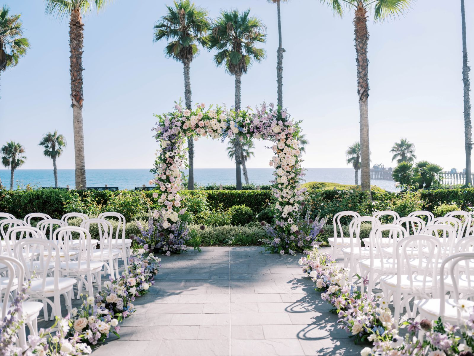Outdoor beachfront wedding setup with floral arch, white chairs, palm trees, and ocean view in background