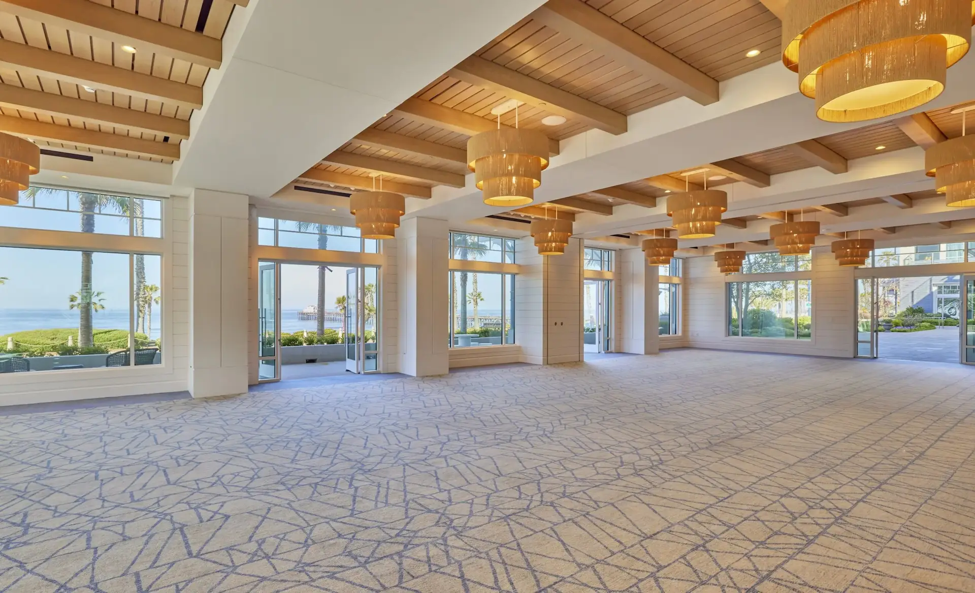Spacious event room with floor-to-ceiling windows, ocean and garden views, wooden beam ceiling, and modern pendant lighting.