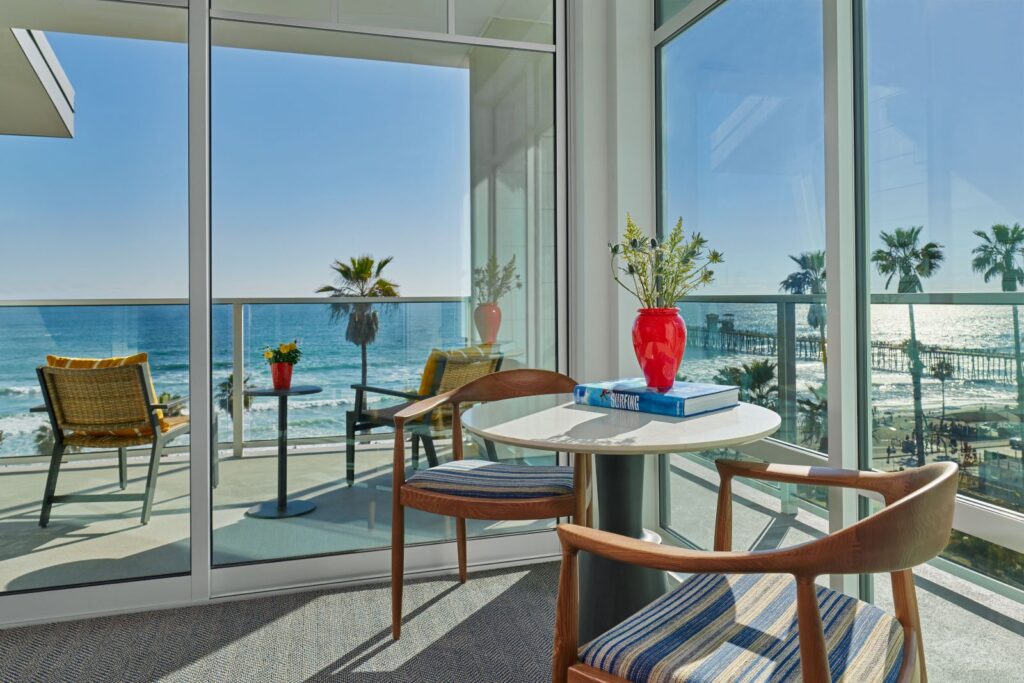 Bright guest room seating area at The Seabird Ocean Resort & Spa with floor-to-ceiling windows, modern wood chairs, and a balcony overlooking the ocean, palm trees, and pier.